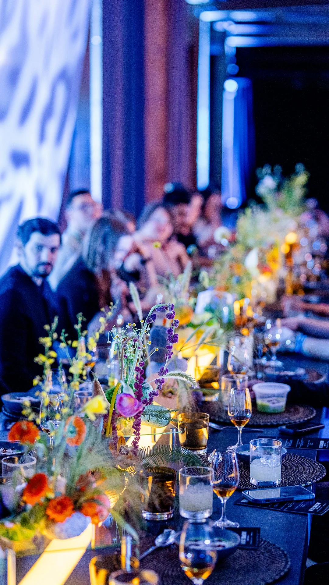 A long dining table set for a formal event with colorful floral centerpieces, wine glasses, and place settings in a dimly lit room with blue lighting.