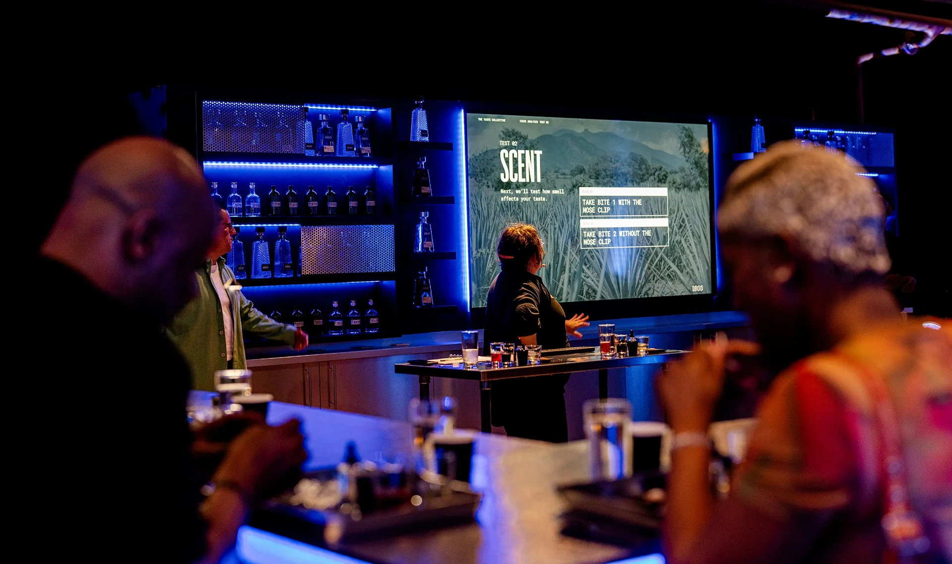 People participating in a scent testing event at a bar or lounge, with a large screen displaying scent options and a row of liquor bottles illuminated in blue behind the bar.