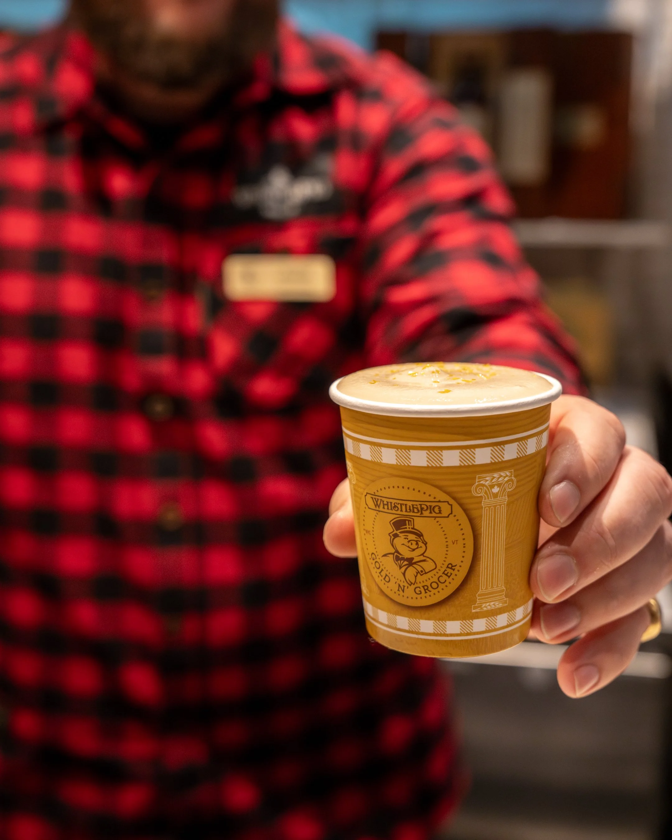 A person in a red and black checkered shirt holding a yellow cup of creamy beverage with citrus zest on top, in a grocery or coffee shop setting.