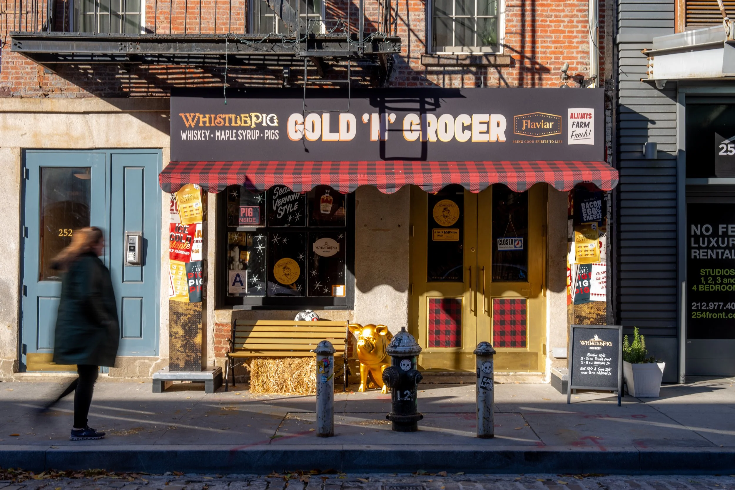 Front view of a storefront named 'Gold 'n' Grocer' with a sign indicating whiskey, maple syrup, pigs, and it has a red and black checkered awning. There is a person walking past the store, a yellow bench, hay bales, and decorative items like a golden pig and a black firefighter statue outside the entrance.