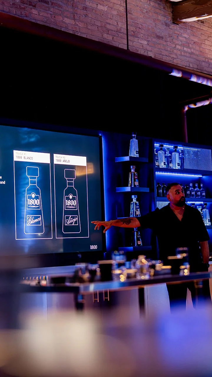 Agave expert, Jaime Salas, pointing at a digital screen with two bottle designs, '1800 Blanco' and '1800 Anejo', in a bar with shelves of liquor bottles illuminated by blue lighting.