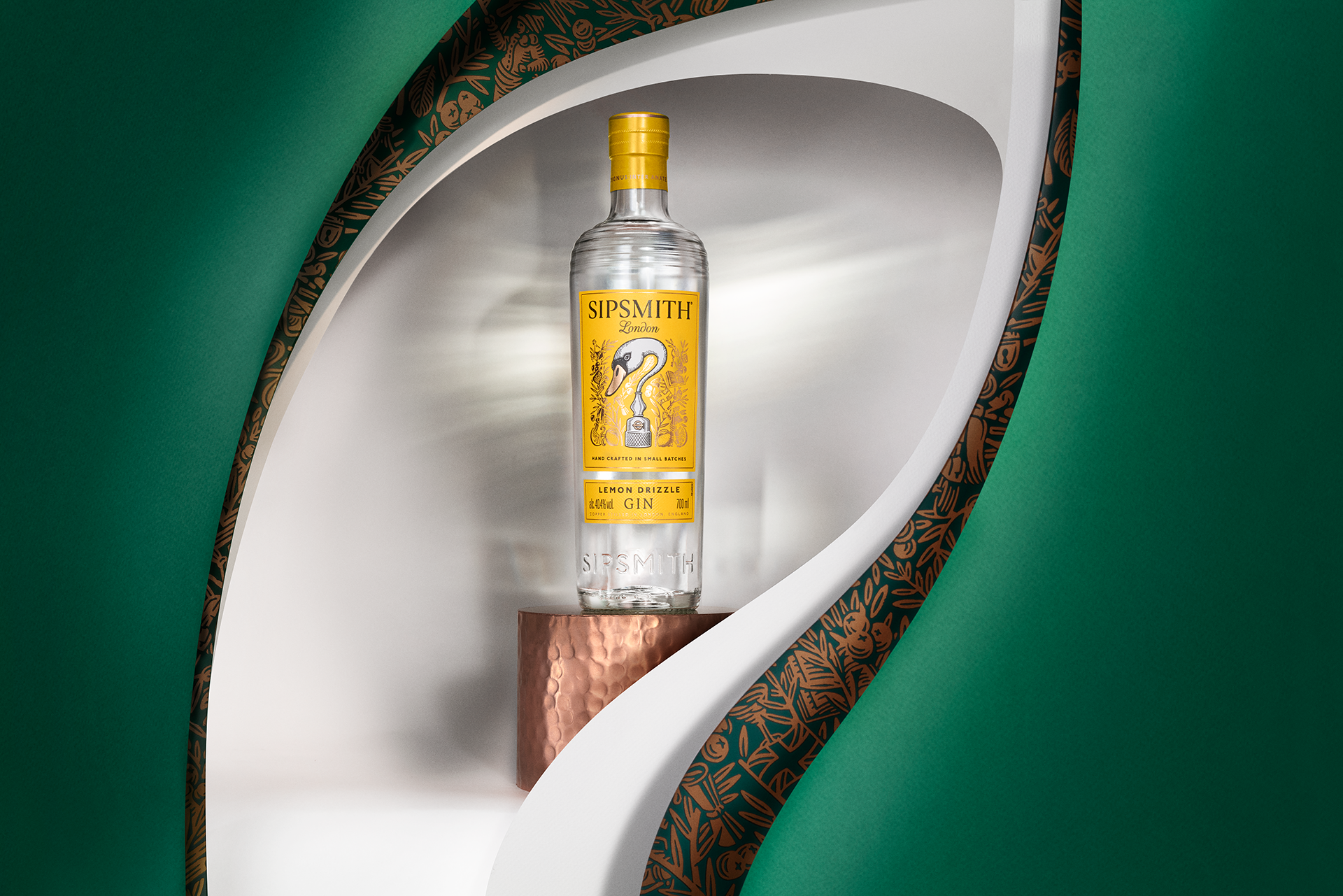 A clear glass bottle of Sipsmith London Lemon Drizzle gin displayed on a small, round, metallic stand inside a white curved niche with green and brown patterned framing.