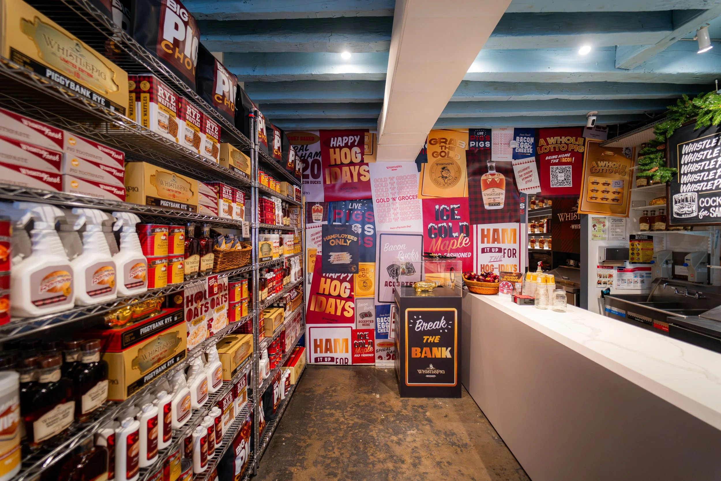 Inside a store decorated with red, white, and black themed signage and banners promoting pork products such as ham, bacon, and sausage. Shelves are stocked with various packaged food products, and a counter with a small basket of apples and some jars is visible. The ceiling is painted in a rustic blue color.