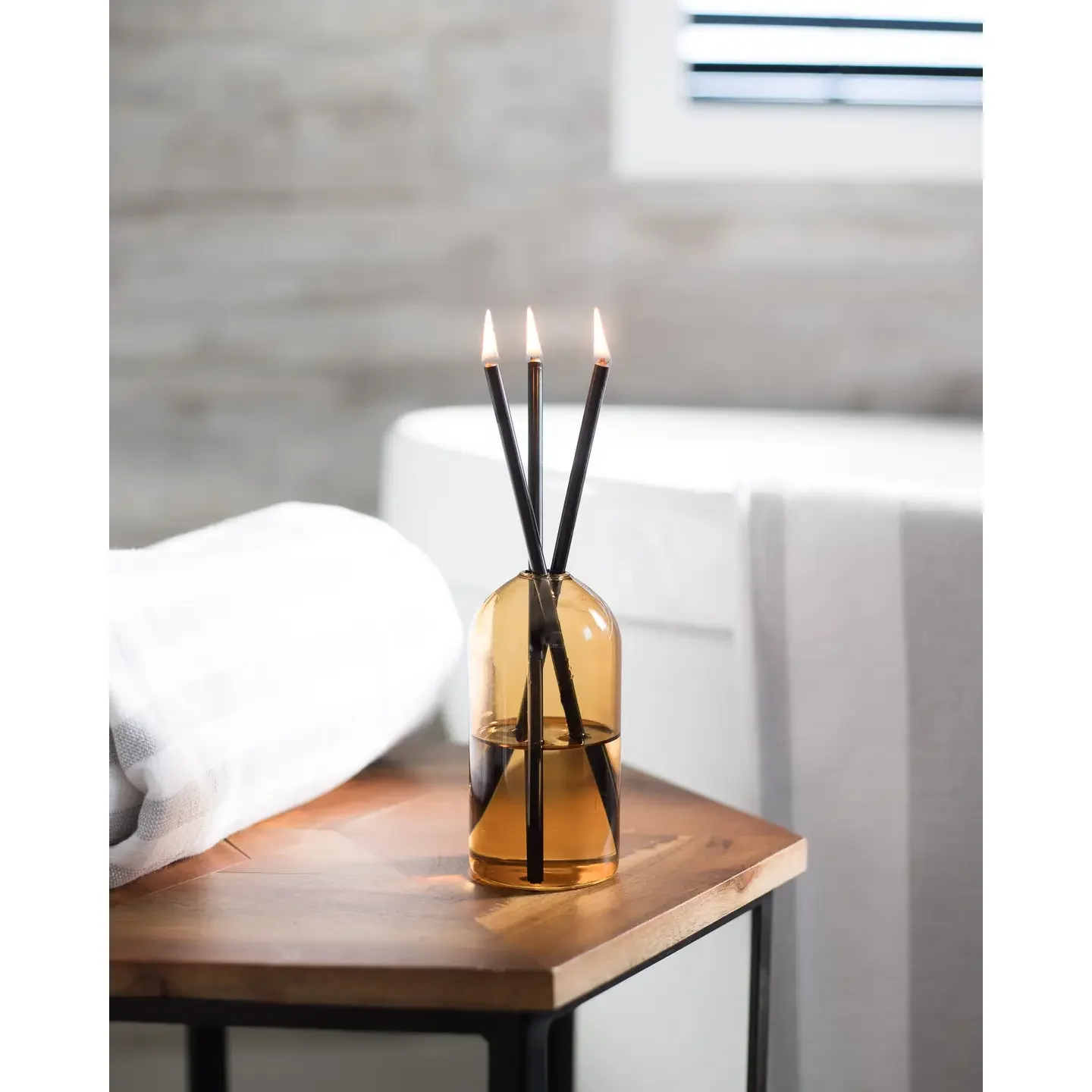 A glass vase with three lit incense sticks placed on a wooden table in a room with neutral walls and a window with blinds.