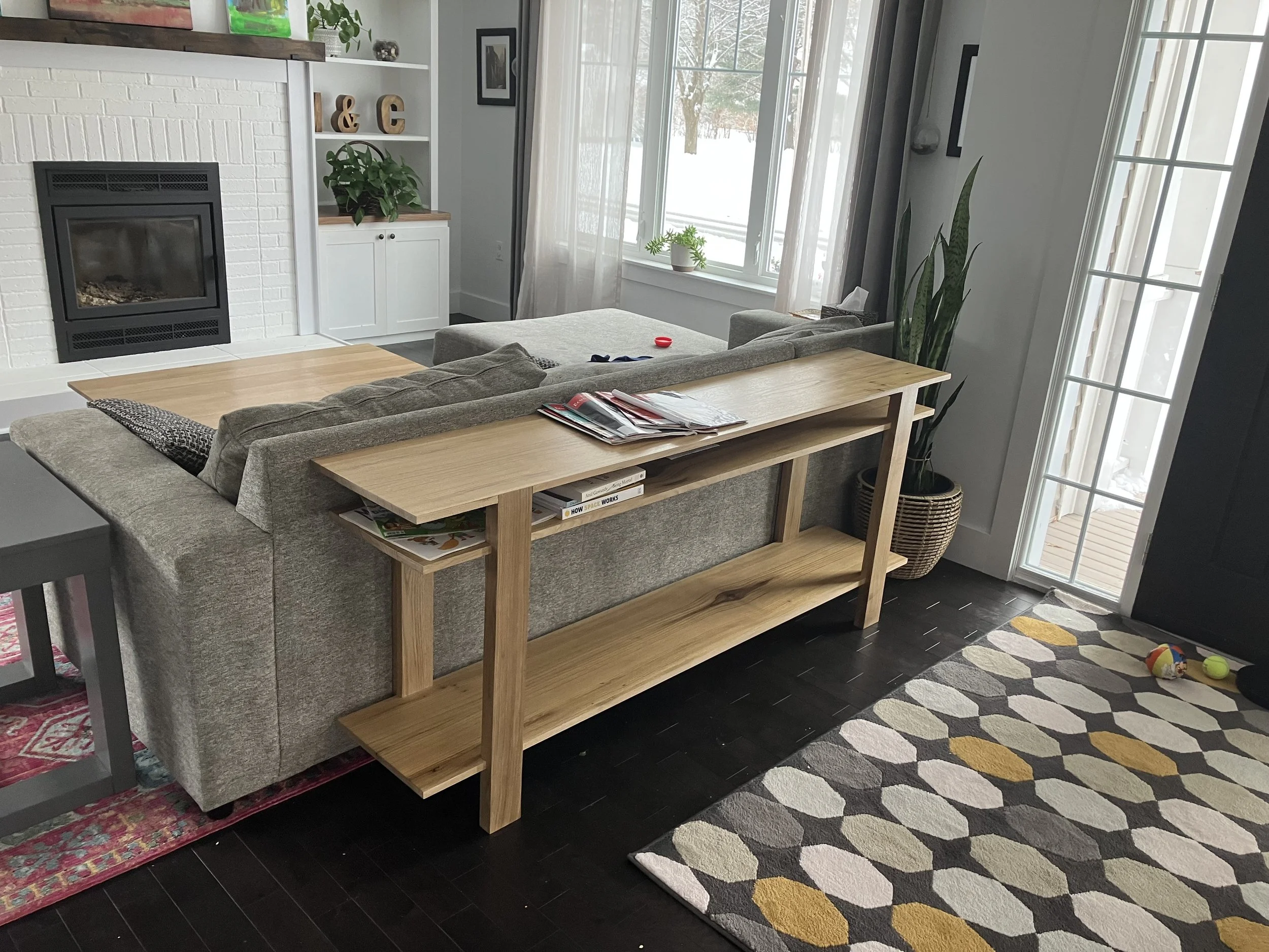 Living room with gray couch, wooden console table, fireplace, window with curtain, potted plant, and indoor toys