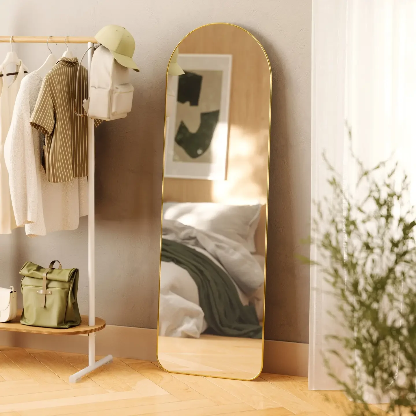 A full-length mirror with a gold frame leaning against a wall in a bedroom. A bed with white sheets and a green blanket is reflected in the mirror. To the left, a clothing rack with beige and striped clothing, a hat, and a beige bag are visible. There's a framed artwork on the wall and some greenery in the foreground on the right.