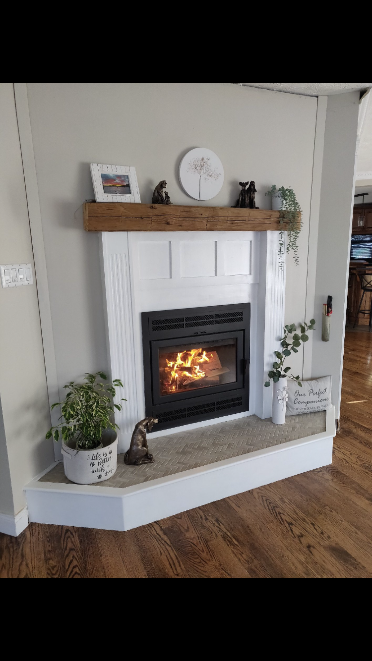 Living room fireplace with white mantel, wooden log shelf, and a fire burning inside. Decor includes plants, framed pictures, animal figurines, and a decorative sign.