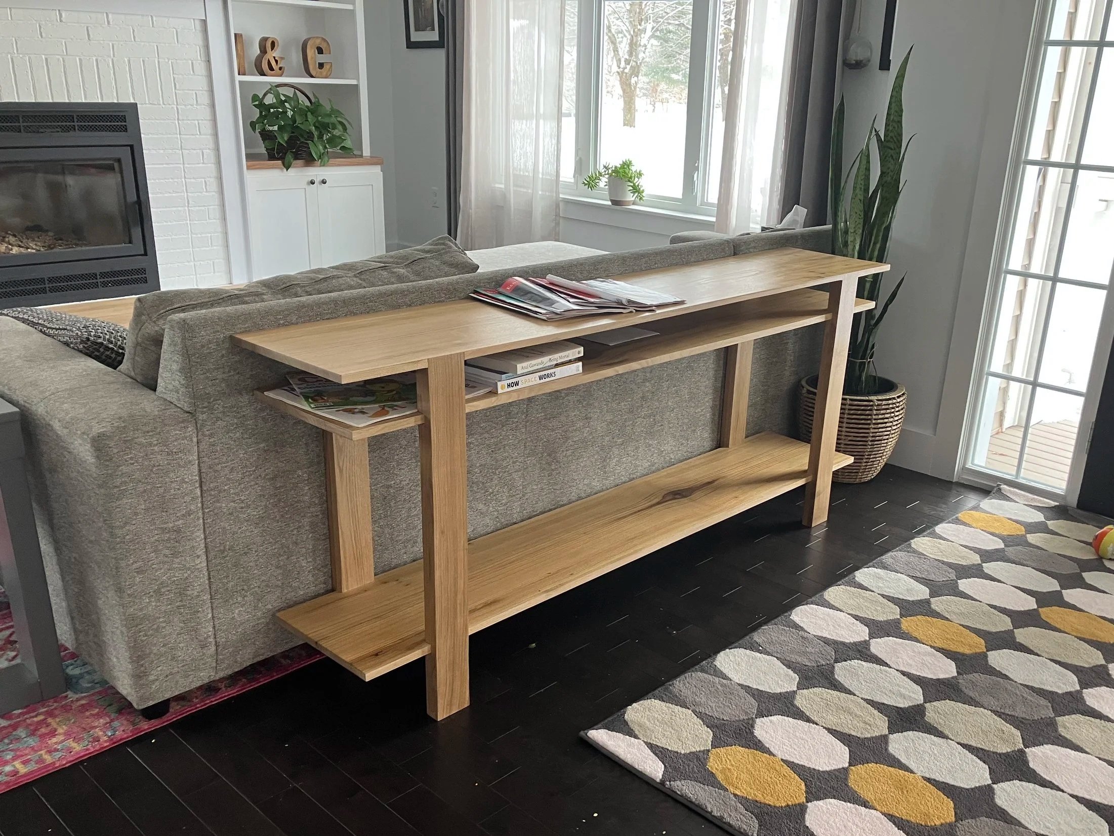 Living room with a beige sectional sofa, a wooden bookshelf behind it containing magazines and books, a window with sheer curtains, a potted plant, a fireplace, a patterned rug, and a glass door leading outside.