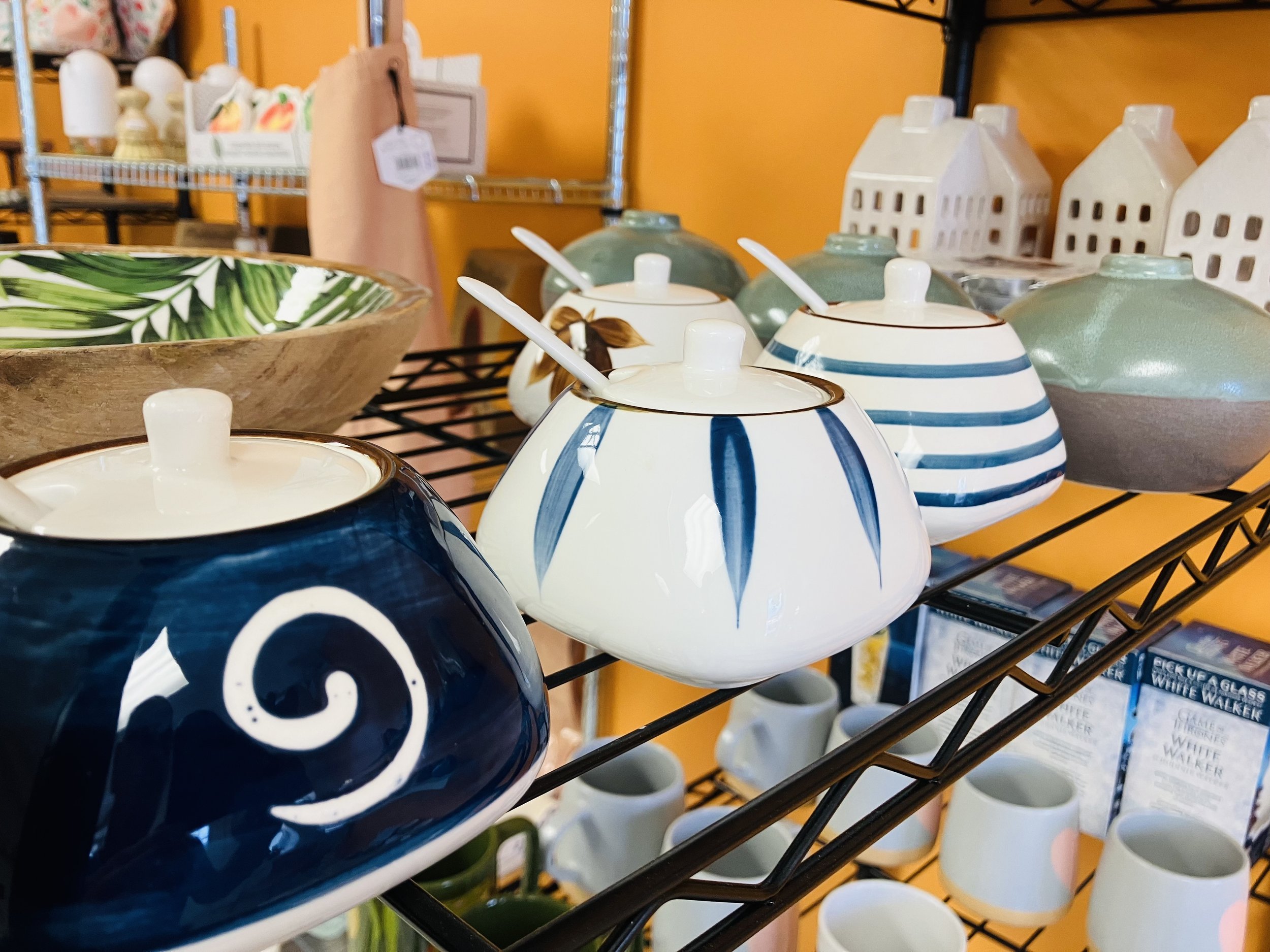 Assorted ceramic sugar bowls and mugs on a black wire shelf in a home decor store, with a yellow wall in the background.
