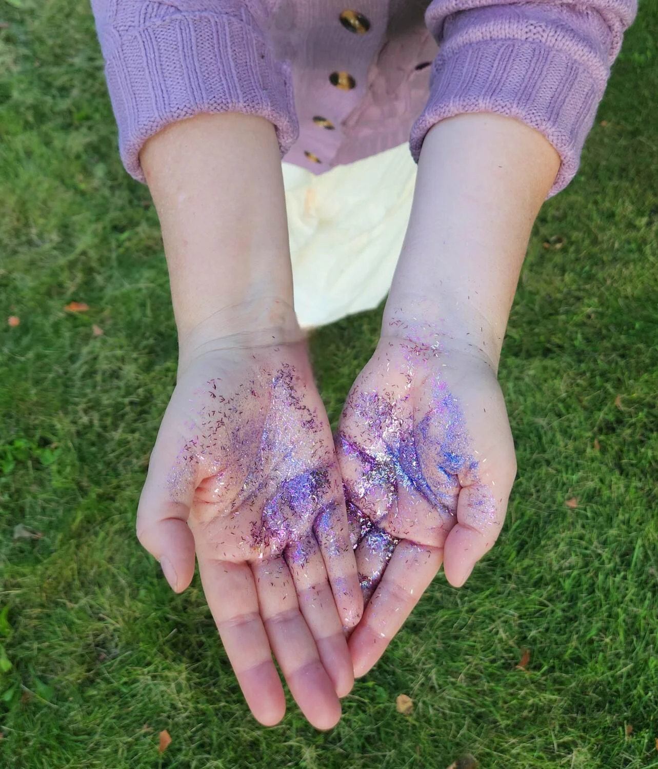 A person's hands covered with purple and silver glitter, held together with palms facing up, outside on green grass.