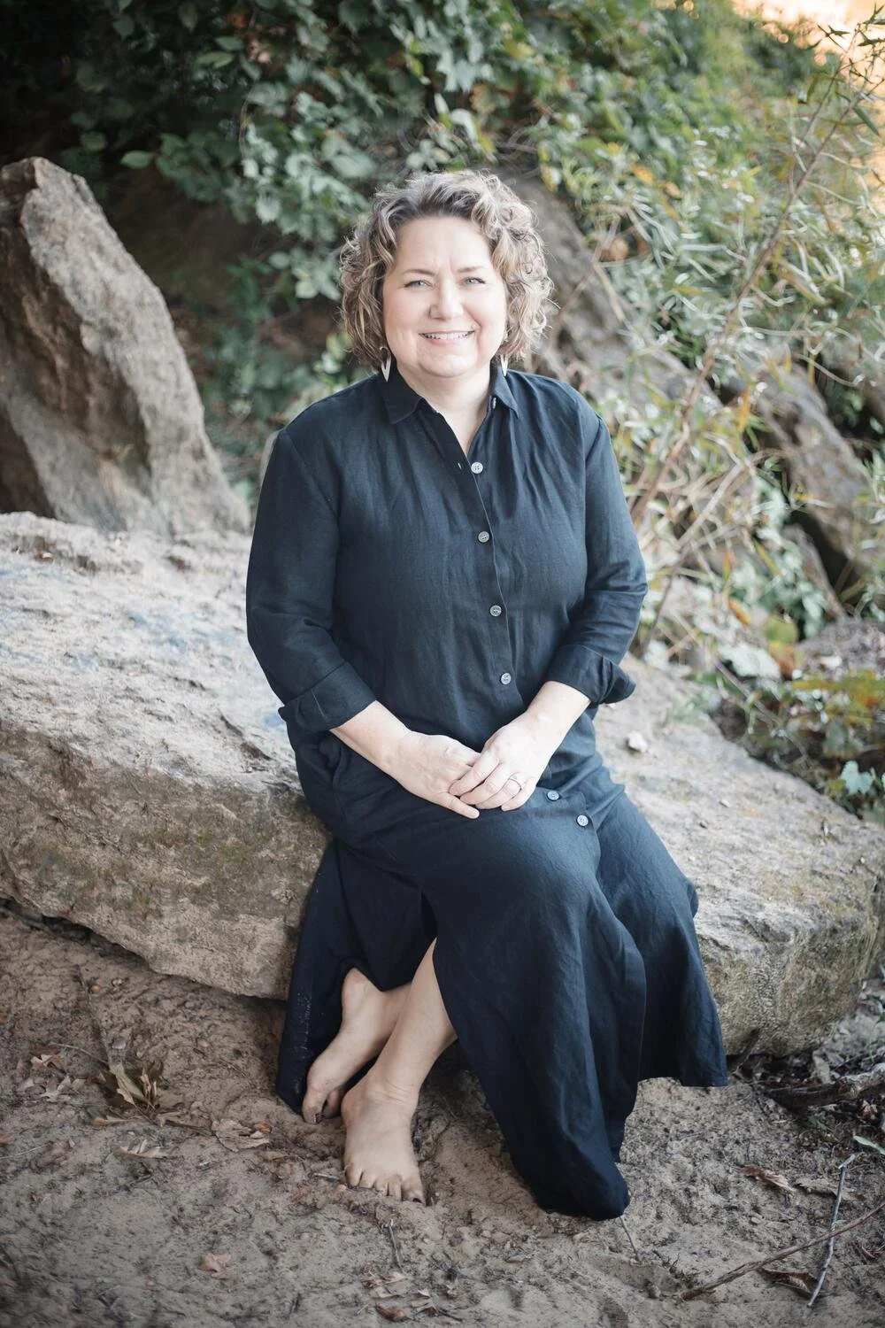 A woman with curly hair and light skin sitting on a large rock outdoors, barefoot, wearing a black dress, surrounded by greenery and rocks, smiling at the camera.