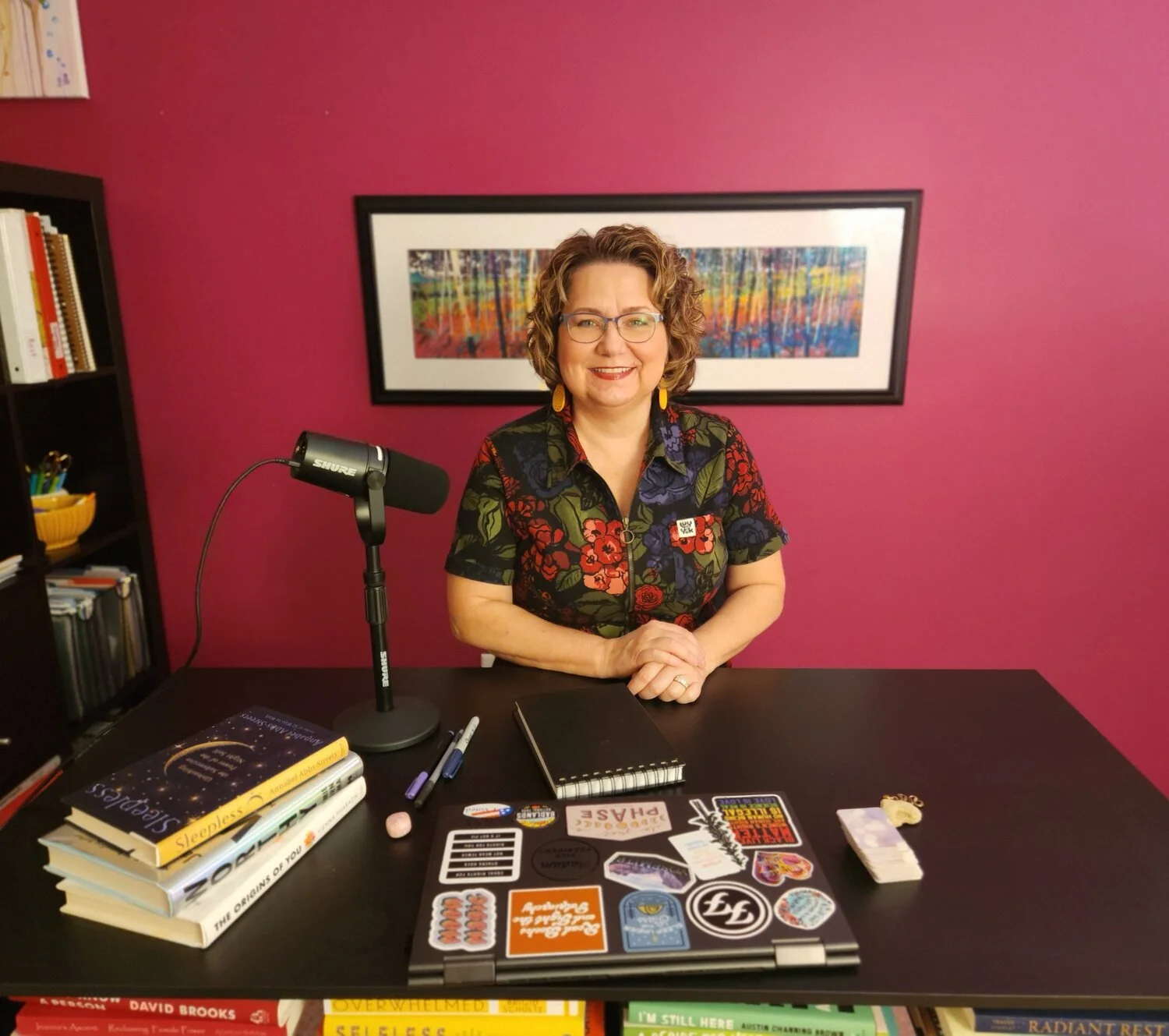 A woman with curly hair and glasses sitting at a desk in front of a pink wall, smiling. The desk has books, a microphone, markers, stickers on a laptop, and a small stack of sticky notes.