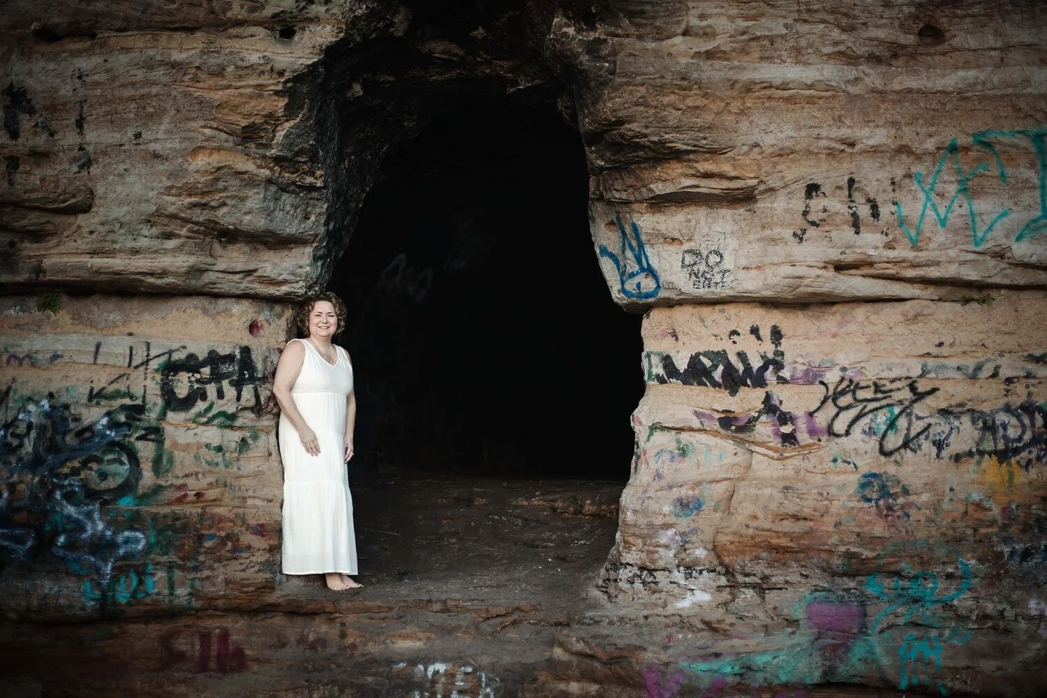 A woman in a white dress standing barefoot in front of a dark cave entrance with graffiti on its rocky walls.