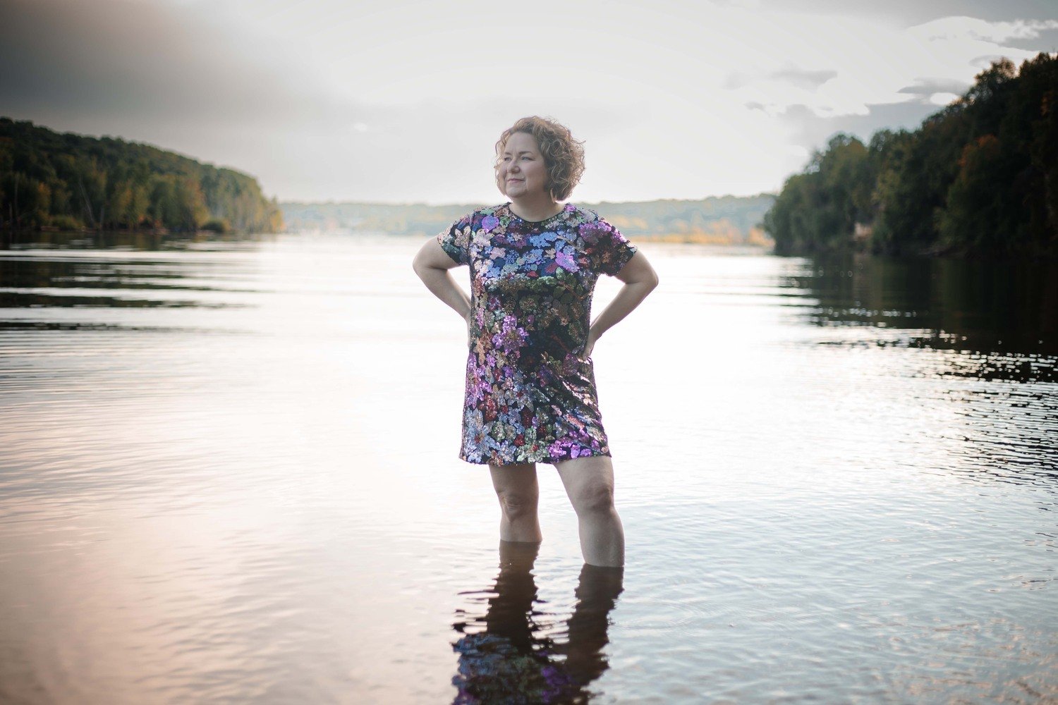 A woman standing in waist-deep water by a river, wearing a colorful floral dress, with her hands on her hips and smiling, with trees in the background and a partly cloudy sky.