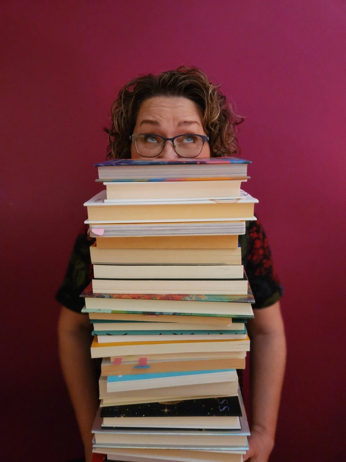 Woman with curly hair and glasses peeking over a tall stack of books against a dark pink background.