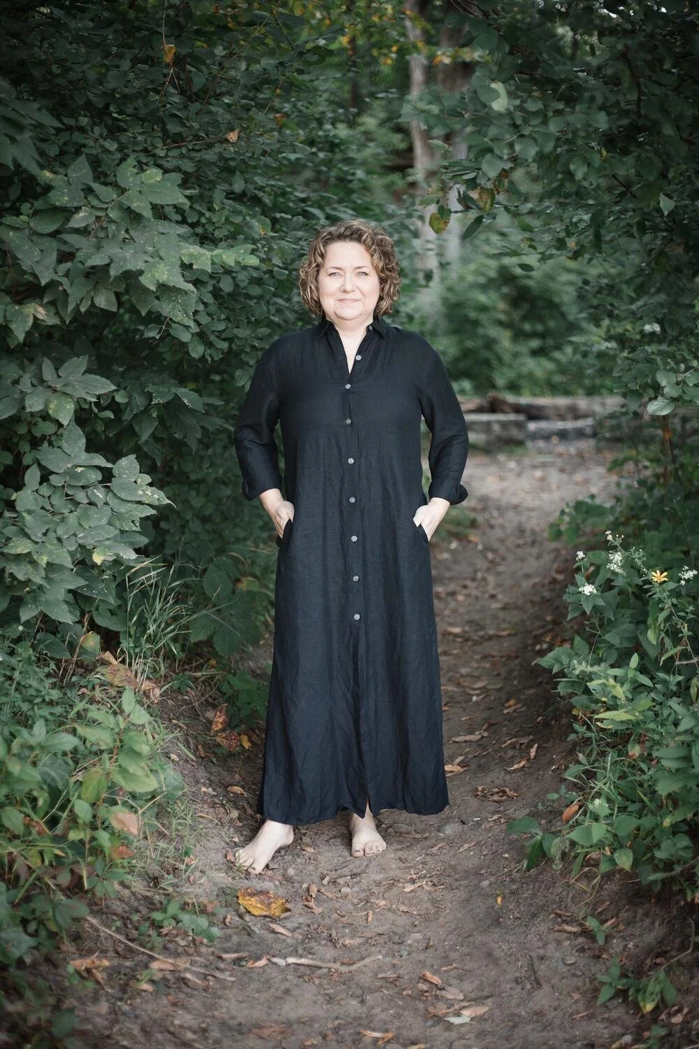 A woman standing barefoot on a dirt trail in a wooded area, wearing a long black button-up dress with her hands in her pockets, surrounded by lush greenery.