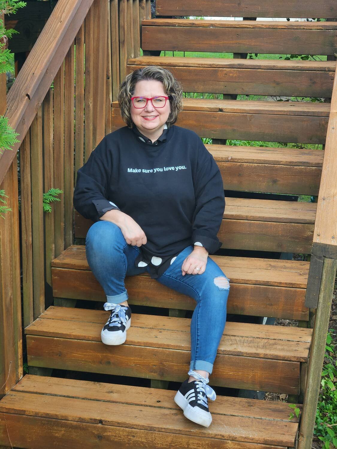 A woman sitting on wooden stairs outdoors, wearing a black sweatshirt with the text "Make sure you love you." and ripped blue jeans, smiling, with red glasses and black sneakers with white stripes.