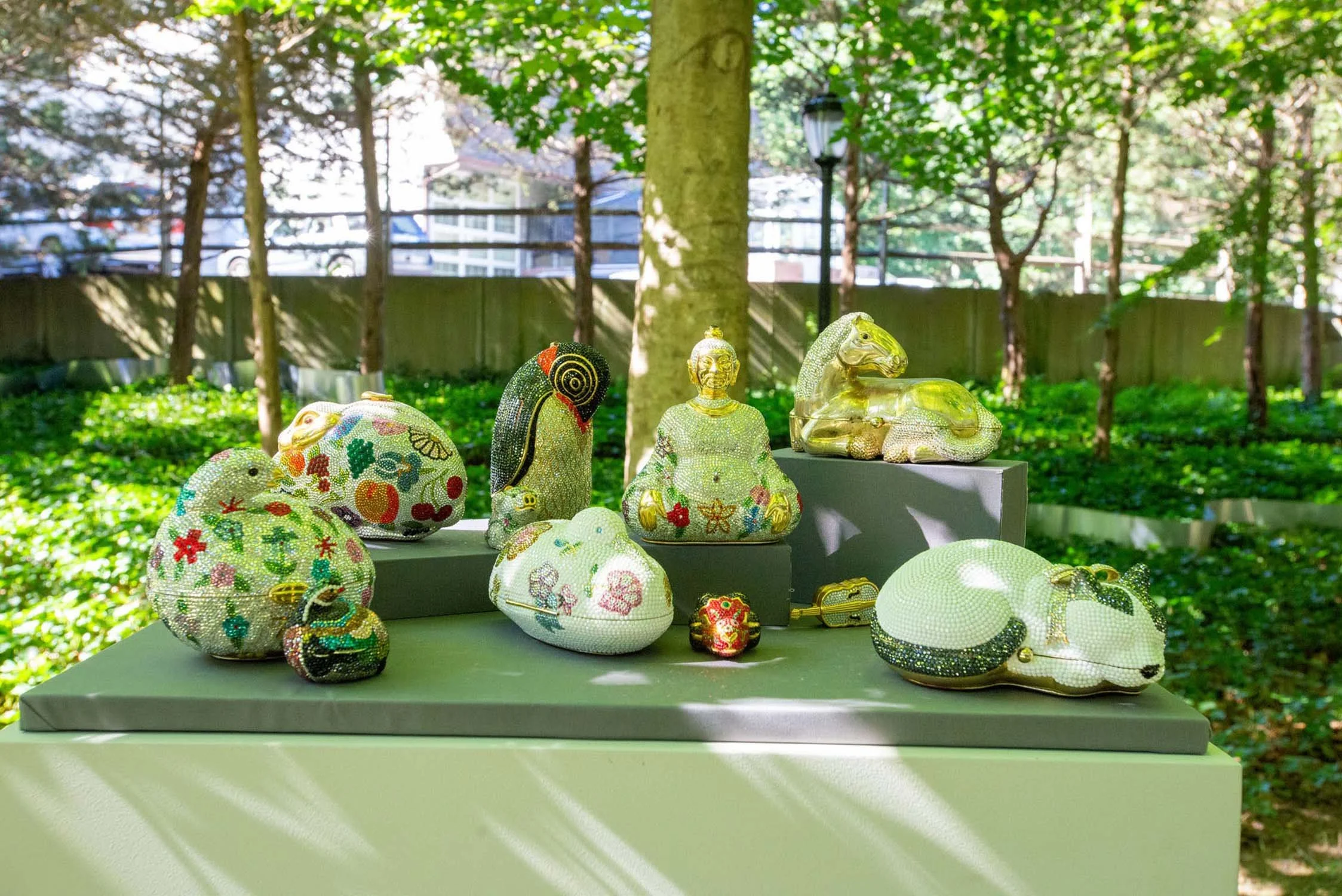 Several decorative animal and figure sculptures covered in colorful beads displayed on grey pedestals outdoors in a green, shaded garden.