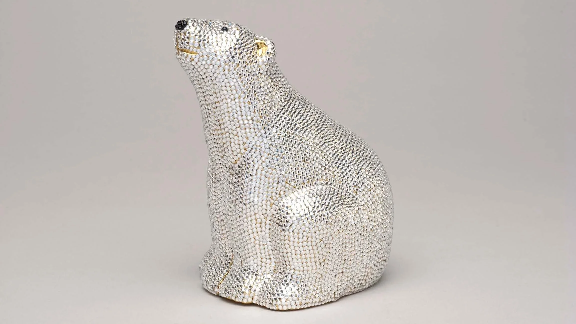A decorative polar bear handbag by Judith Leiber covered in clear rhinestones with gold accents on the nose and eyes, placed on a neutral background.
