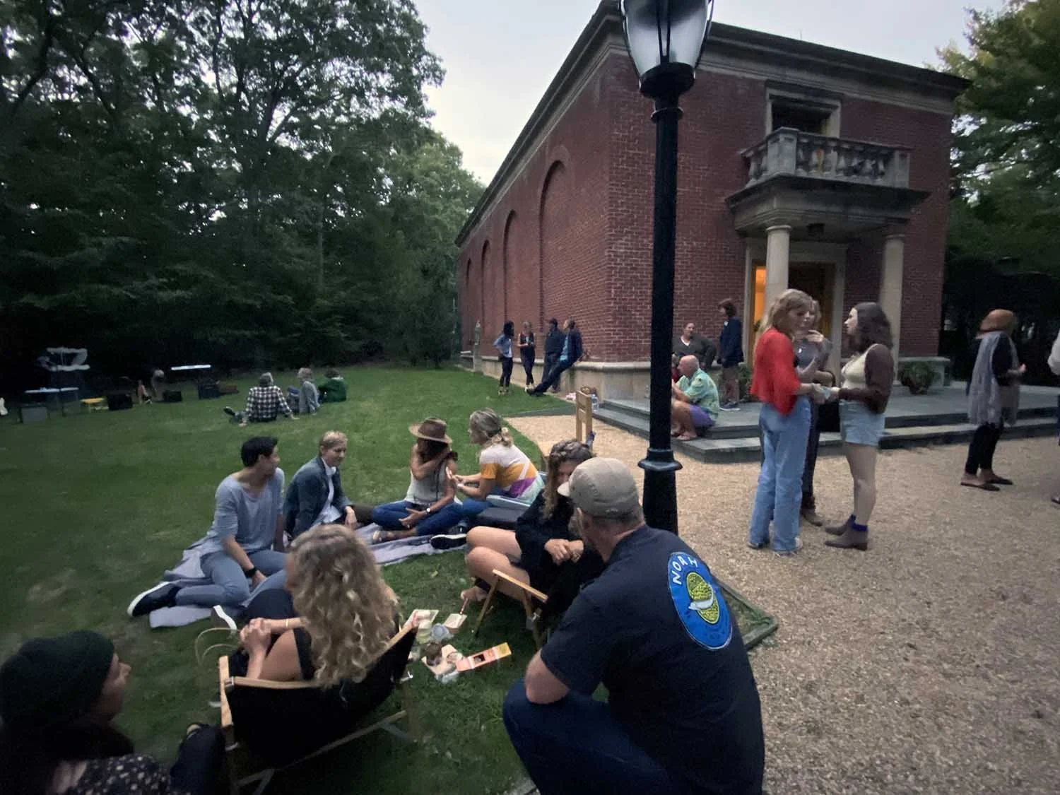 Group of people gathered outside The Leiber Collection museum historic red brick building, socializing on the steps and lawn during the evening, with a lamppost in the foreground and trees in the background.