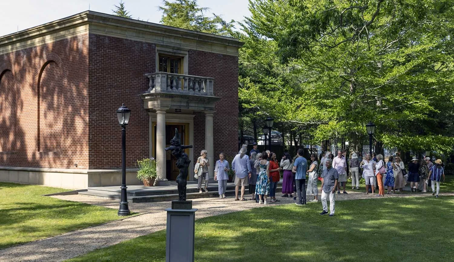 Group of people gathering outside a The Leiber Collection museum brick building with a small balcony, trees, and greenery.