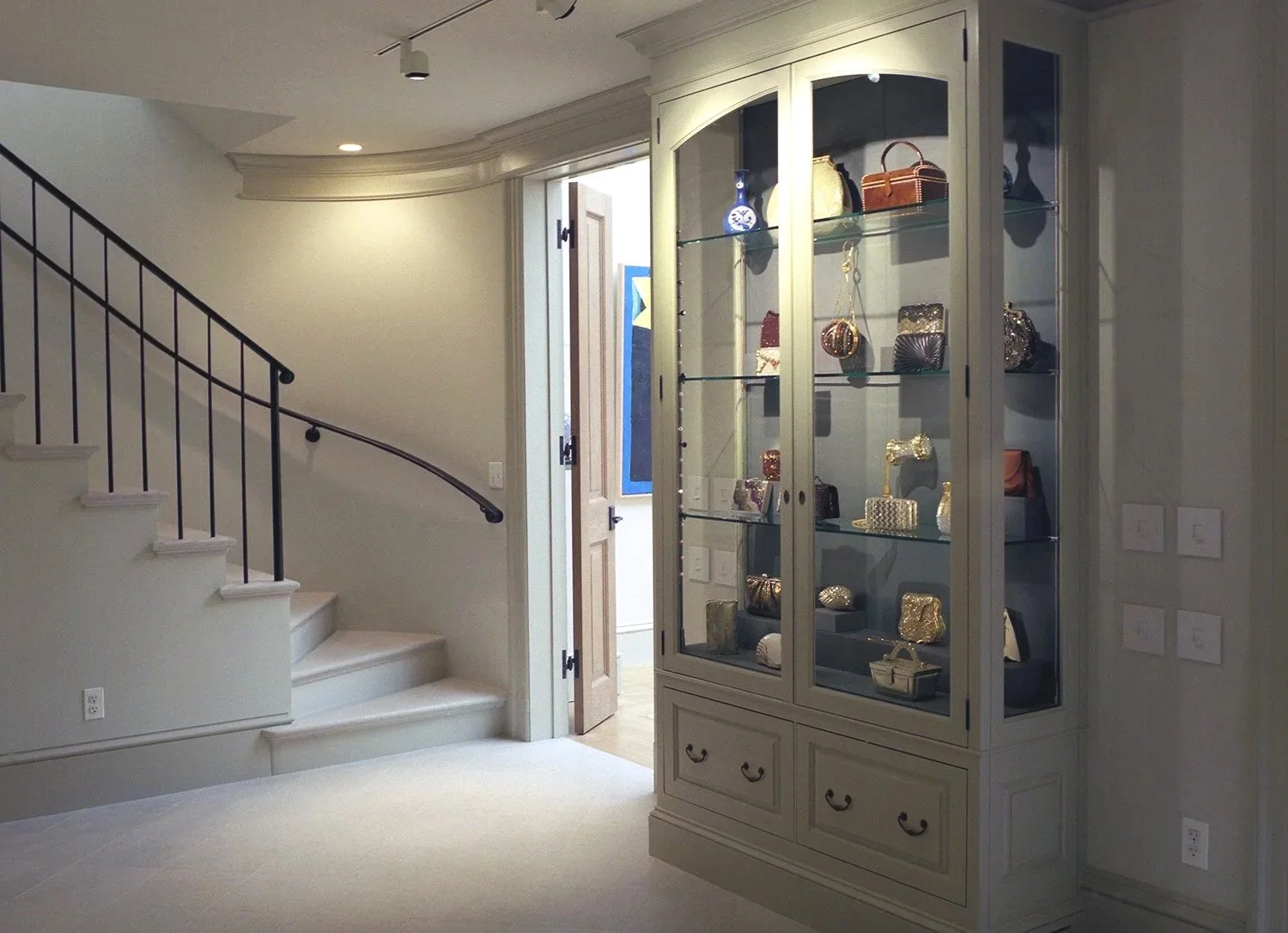 Interior of The Leiber Collection museum with a glass display cabinet showcasing Judith Leiber luxury handbags, a staircase with a black railing, next to an open door leading to another display room.