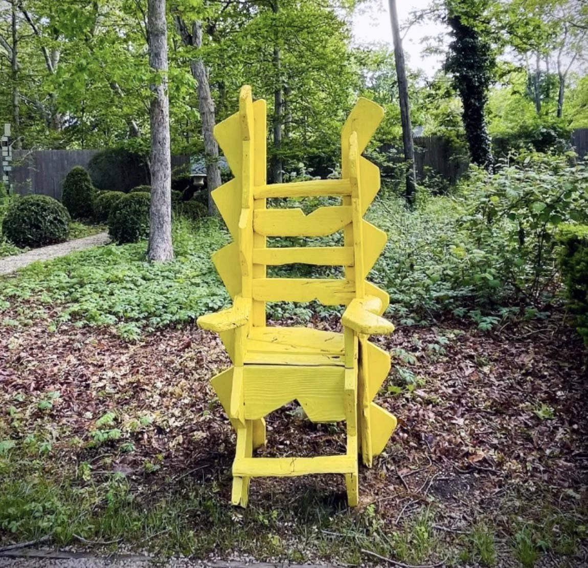 A yellow painted wooden chair placed upside down in a lush green garden surrounded by trees and bushes.