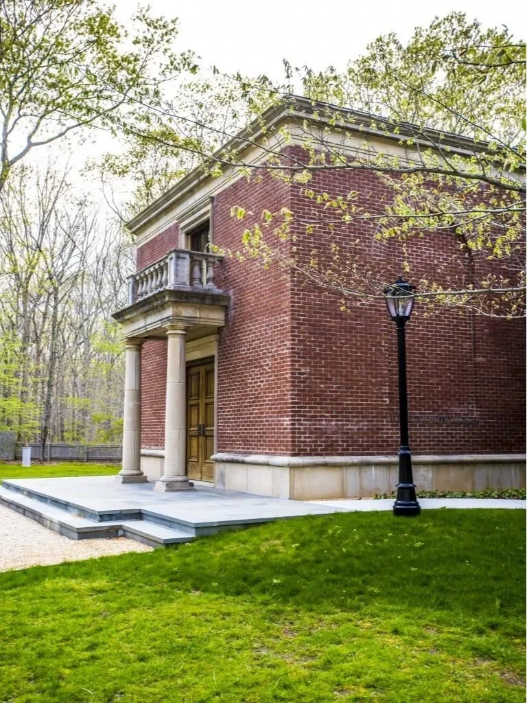Brick building with a small balcony, columned entrance, and a black street lamp in front, surrounded by grass and trees.