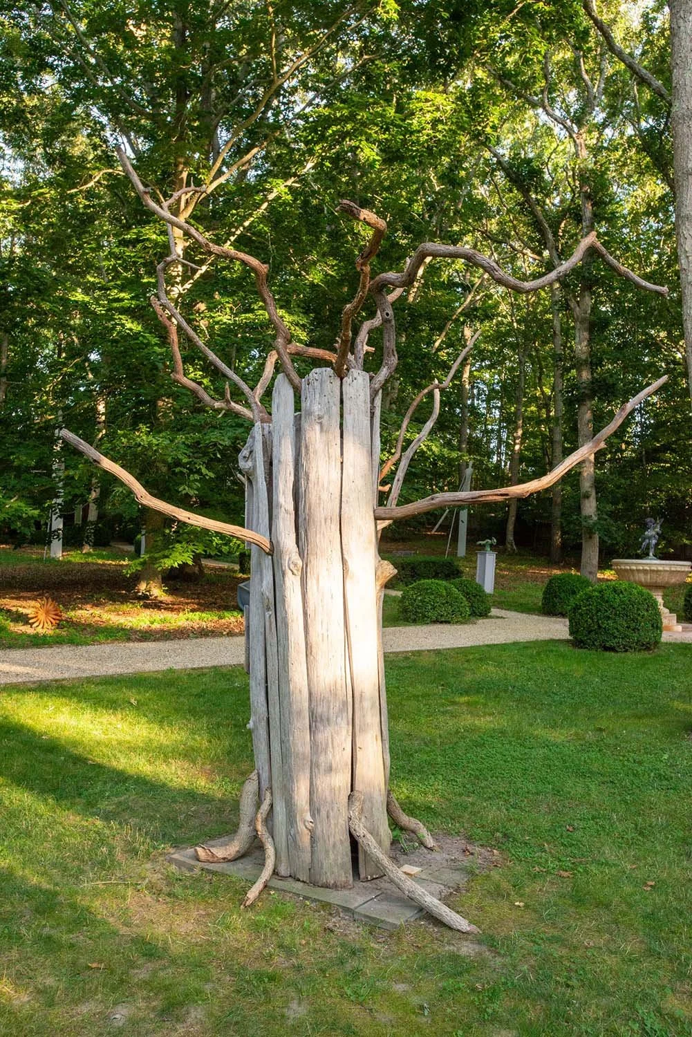 A sculpture of a tree with twisted, leafless branches grows from an old wooden post in a lush green garden.