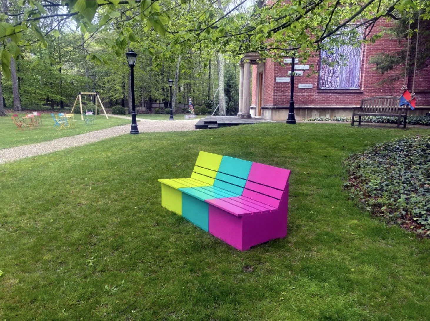 Colorful bench in a green park with a brick building, picnic tables, swings, and lamp posts.