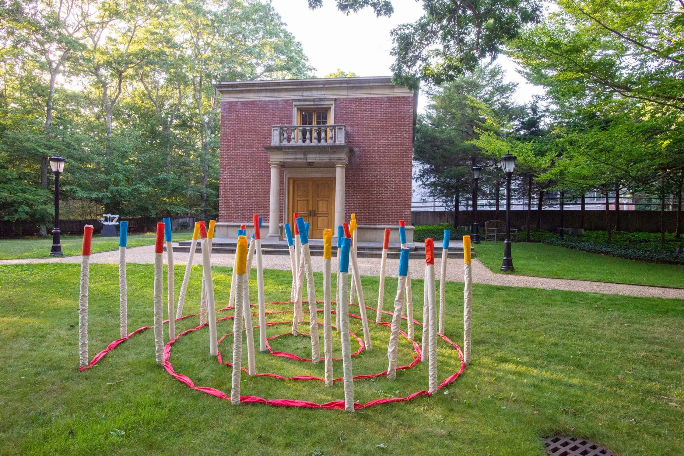The front the Leiber Collection museum, a brick building with a wooden door and small balcony surrounded by green grass. In the foreground, a colorful ring toss game with tall, wrapped sticks with red, blue, and yellow tops inside a pink ring.
