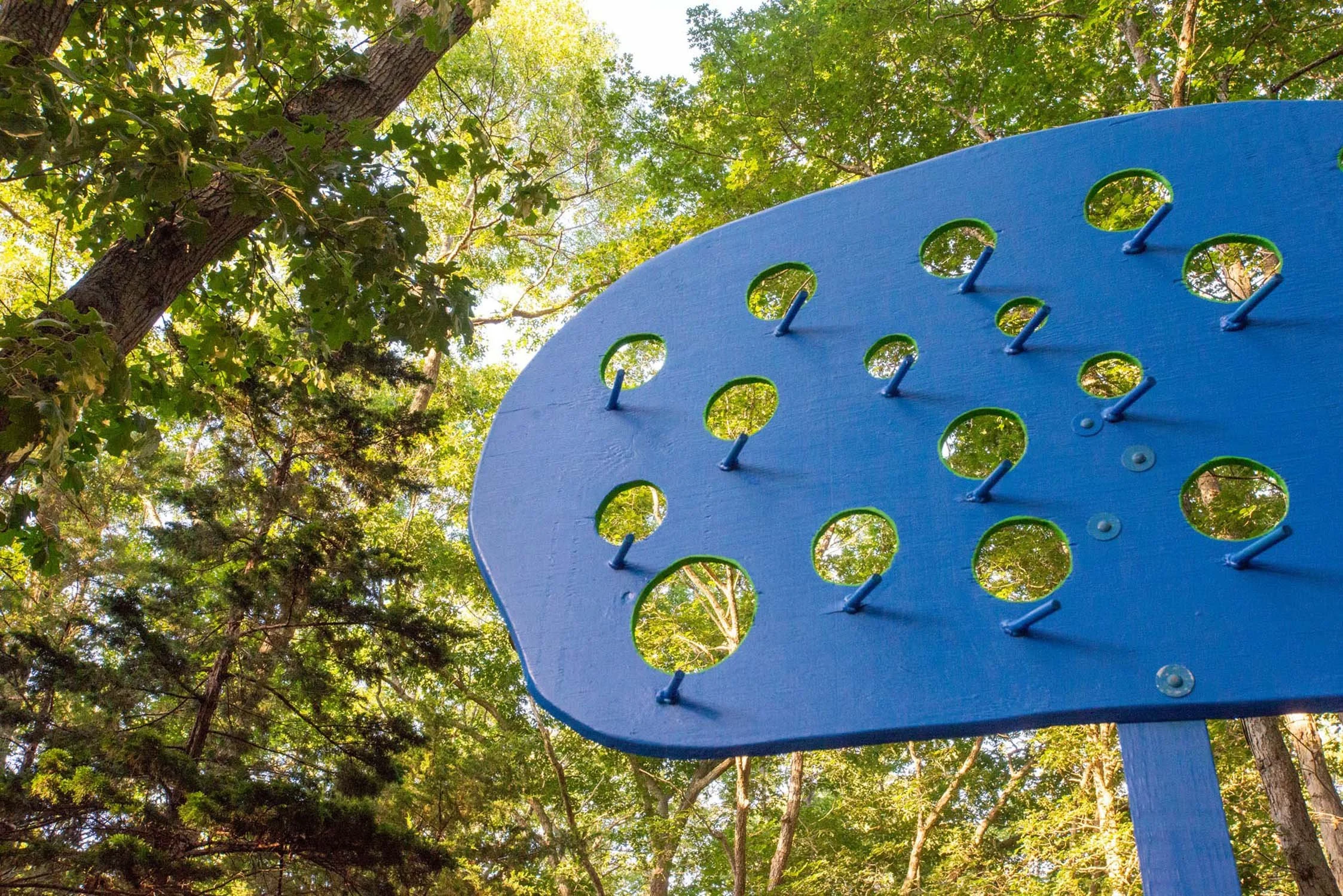 Close-up of a blue sculpture with multiple circular openings, set against a background of tall trees and green leaves.
