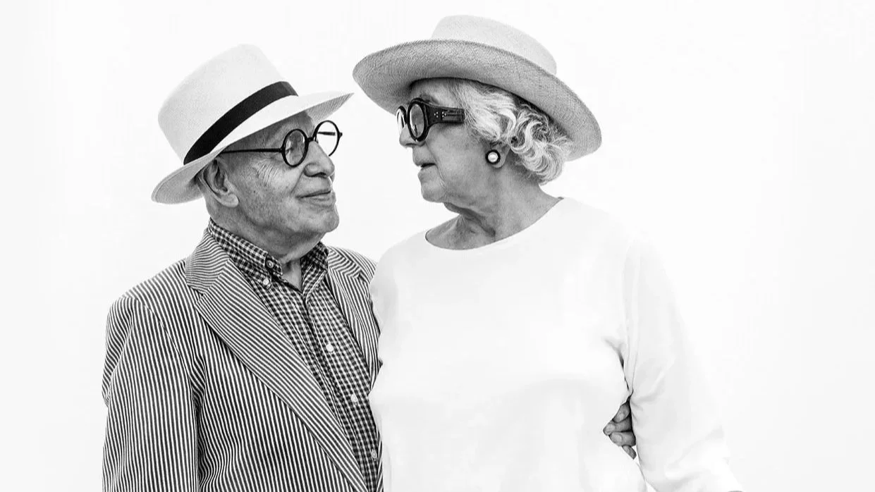 Gerson and Judith Leiber in vintage clothing and hats, looking at each other closely, standing against a plain white background.