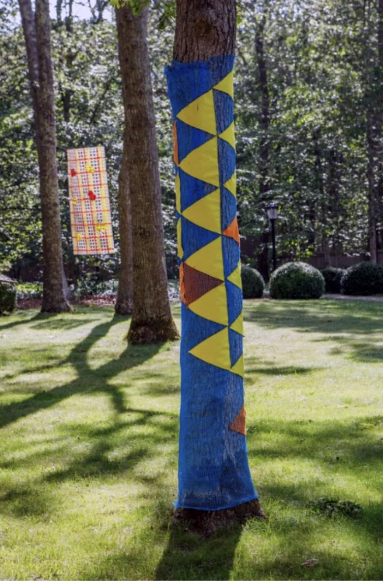 Colorful outdoor sculpture wrapped around trees in the garden, with one tree painted blue with yellow and orange geometric shapes, and another piece in the background featuring a red and white checkered pattern with small decorative elements.