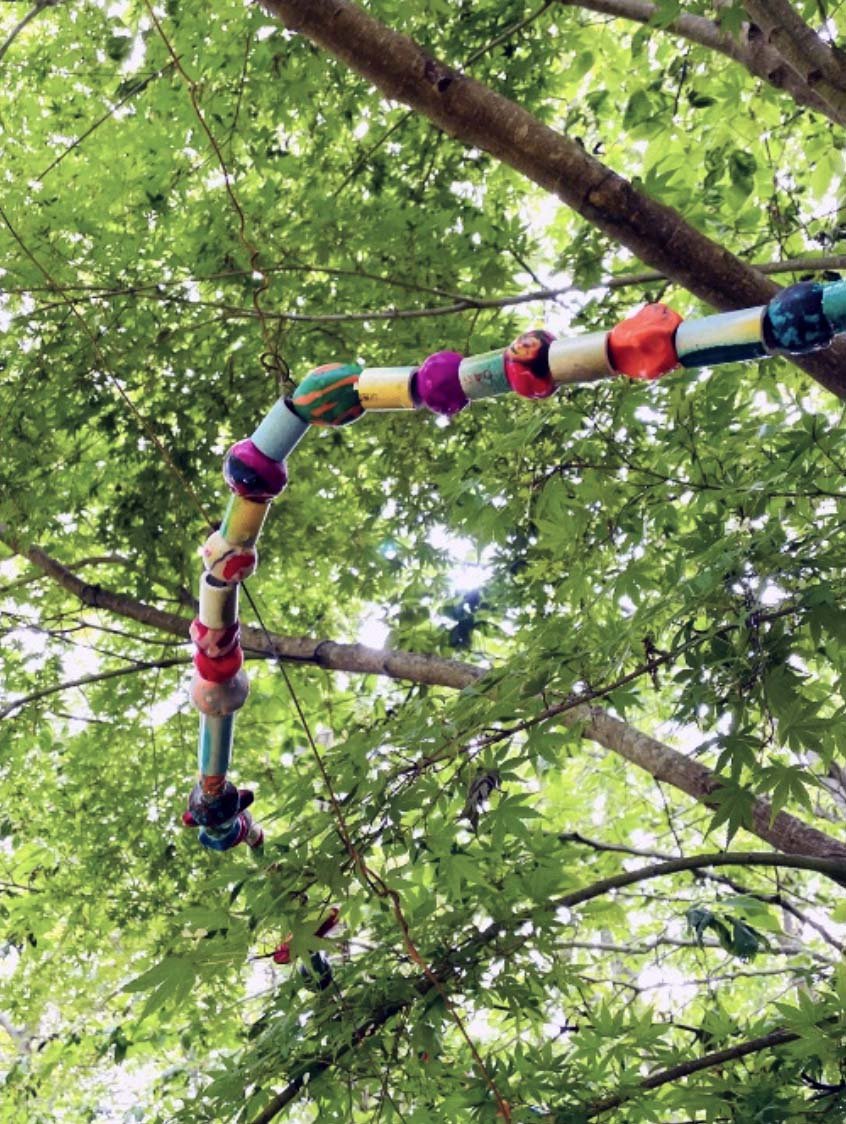 Colorful decorative sculpture consisting of connected spheres and cylinders hanging from tree branches with green leaves overhead.