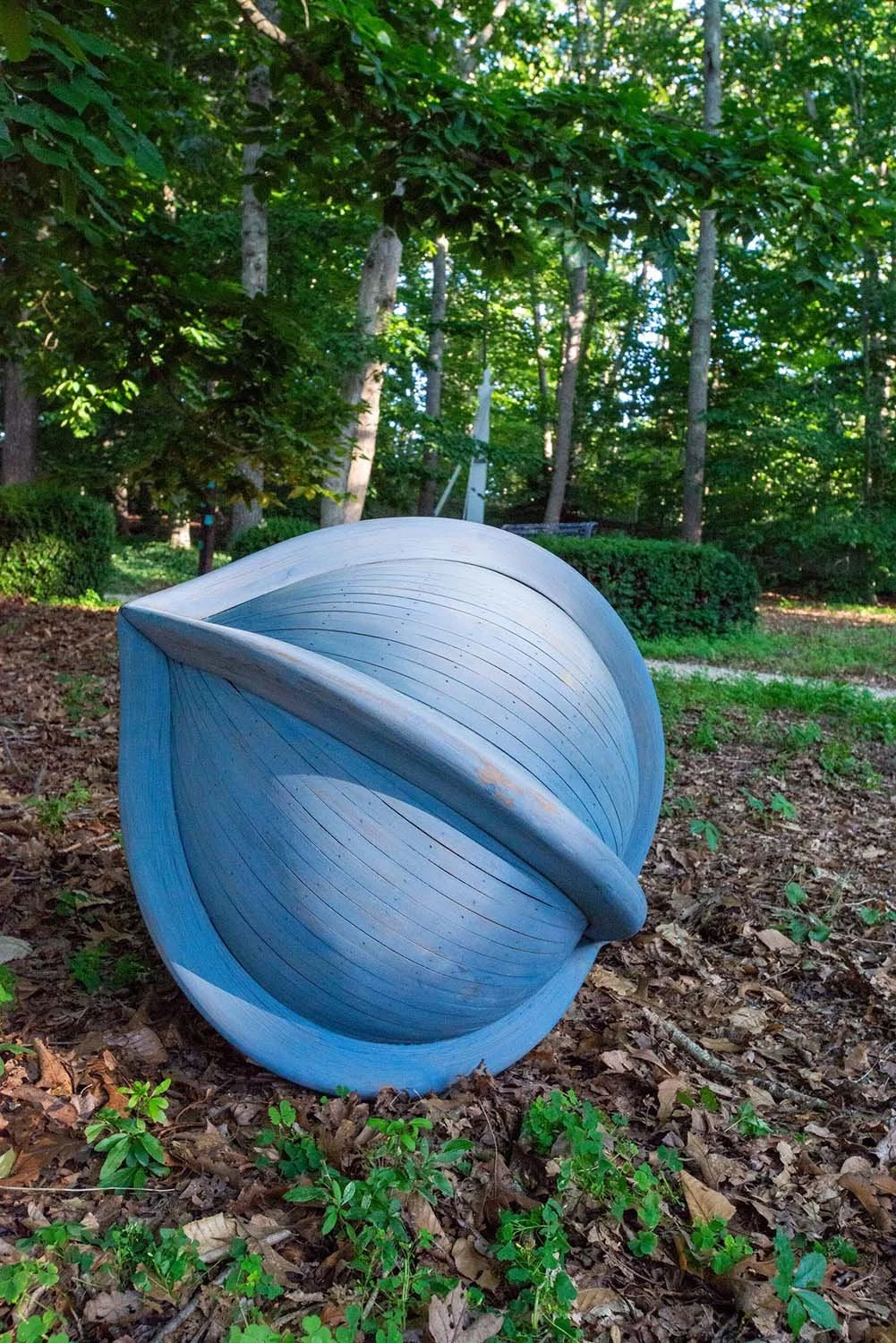 Blue sculpture resembling a planet with a ring, situated in a wooded outdoor area with trees and greenery.