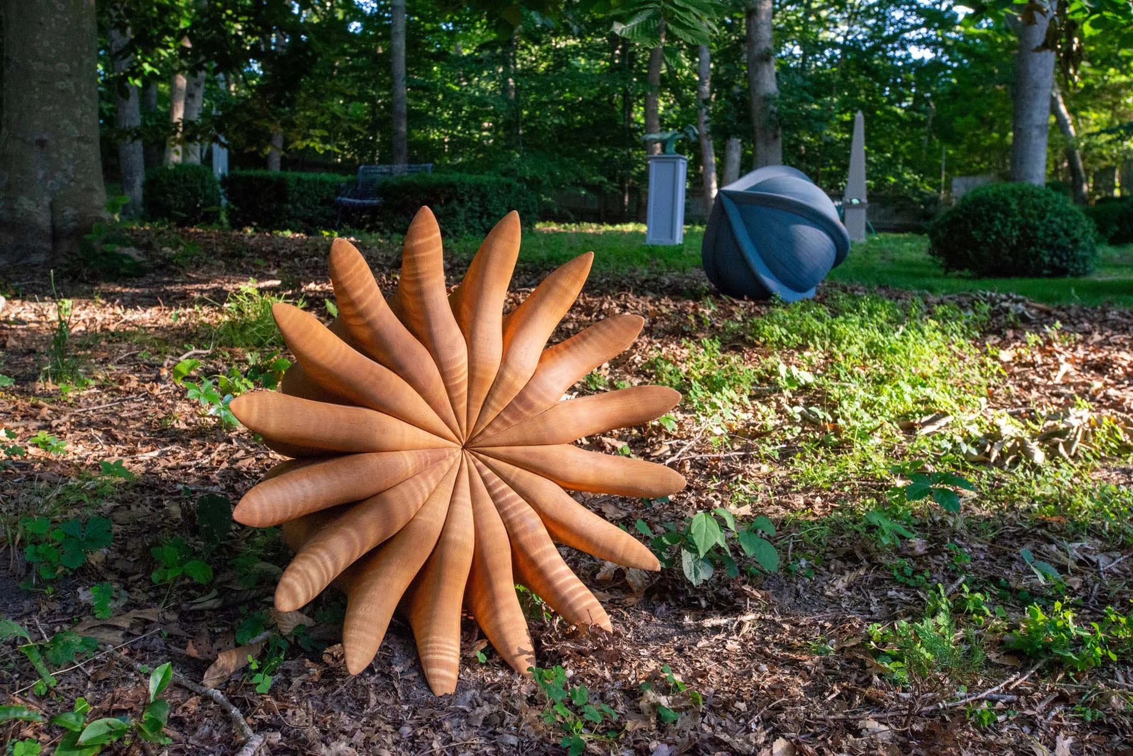 Outdoor garden scene with a large wooden sculpture resembling a flower in the foreground, surrounded by green plants, trees, and some outdoor furniture in the background.