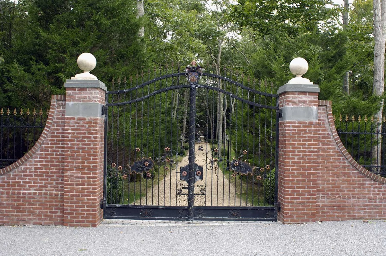 Decorative wrought iron gate at The Leiber Collection entrance with floral and butterfly motifs, flanked by brick pillars topped with white spherical ornaments, leading into a garden pathway surrounded by lush green trees.