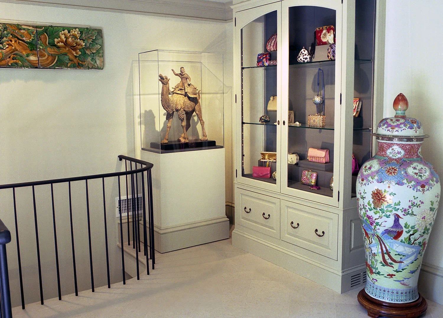 Interior room of The Leiber Collection with a large painted porcelain vase, a display case with small elegant, whimsical handbags by Judith Leiber, a carved wooden sculpture of a camel with a rider, and a colorful wall art piece.