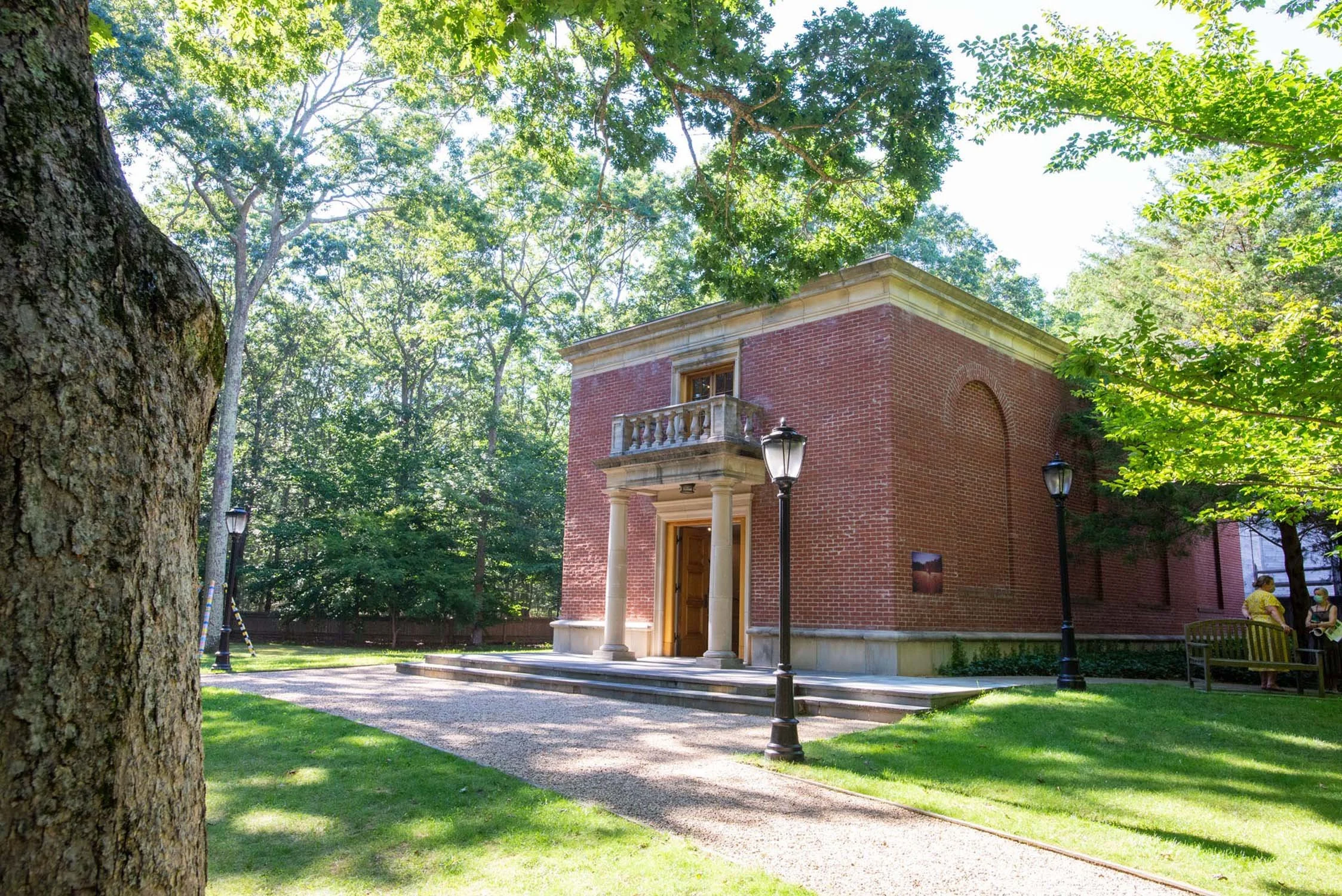 The Leiber Collection museum historic brick building with columns and a small balcony, surrounded by trees and greenery, with a gravel pathway and vintage-style lamp posts in the front.