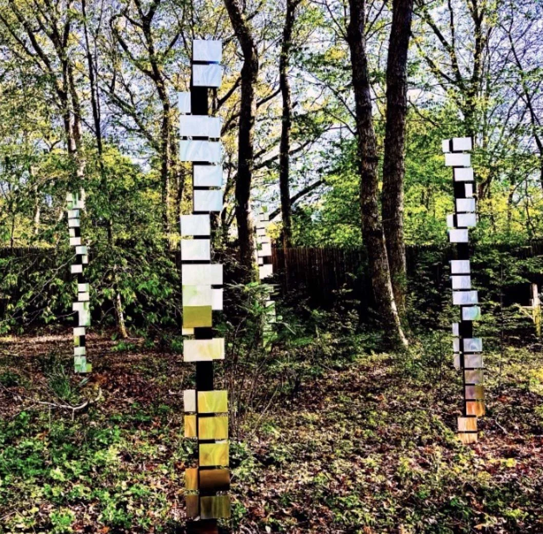 Outdoor art installation of three tall vertical structures made of rectangular reflective panels arranged in a spiral pattern in a wooded area with trees and green foliage.