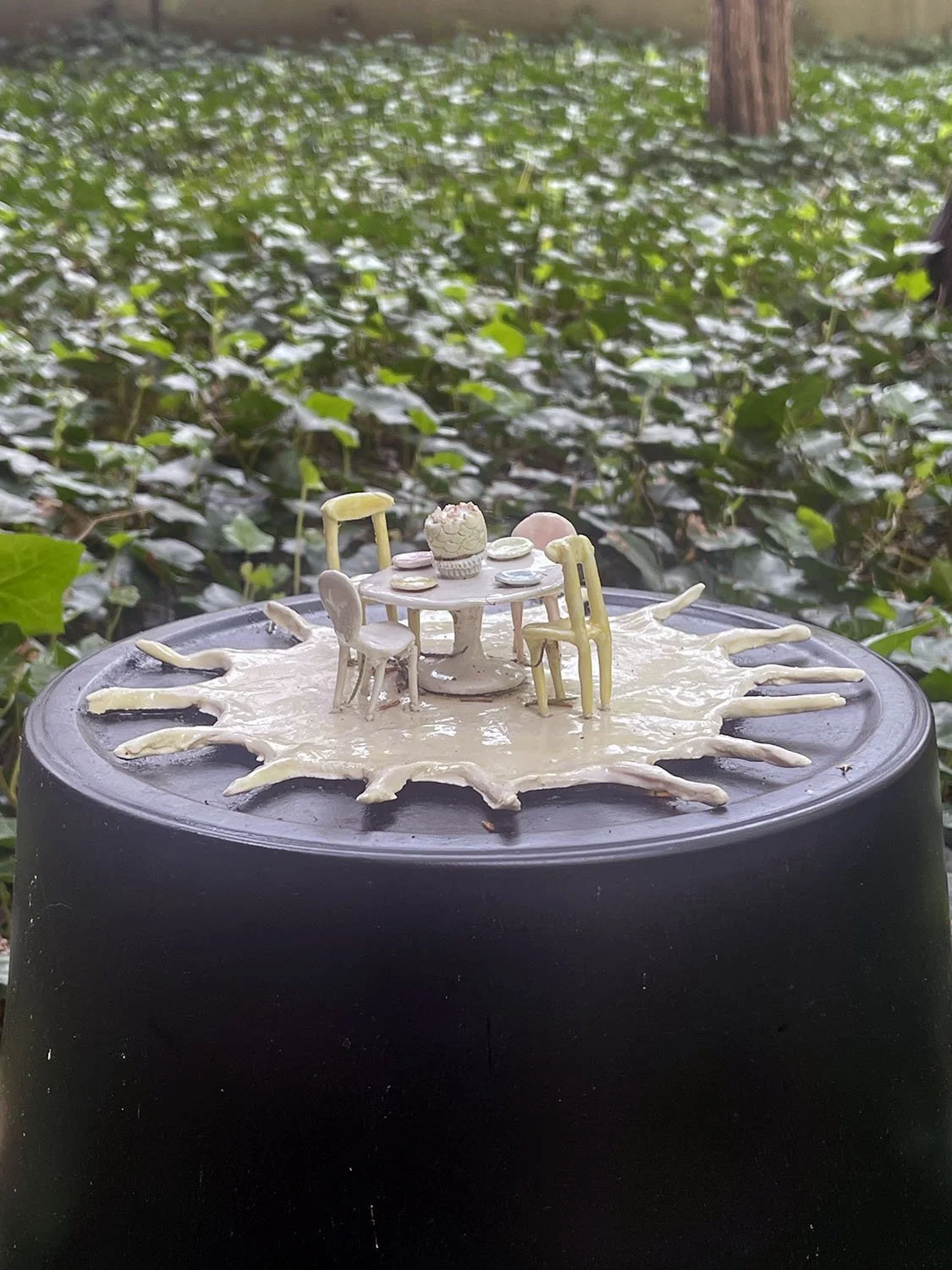 Miniature dollhouse furniture table and chairs with a cake on top, set on an outdoor surface with ivy and trees in the background.