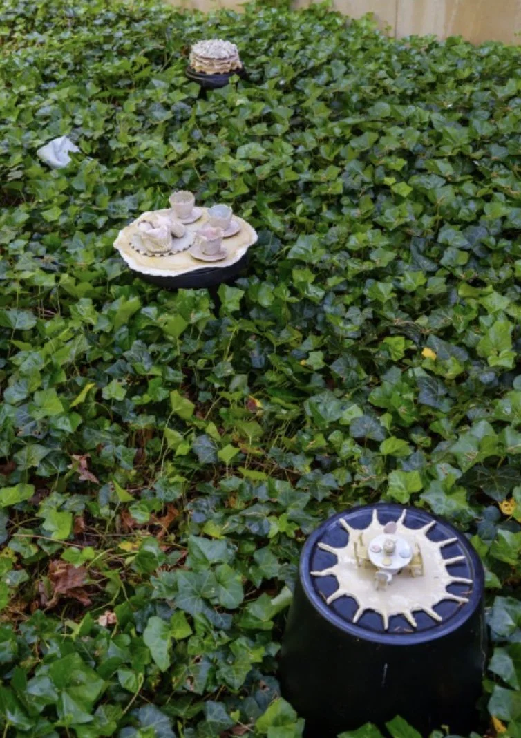Three black cylindrical tables with miniature tea sets on lace doilies placed in a garden surrounded by green ivy plants.