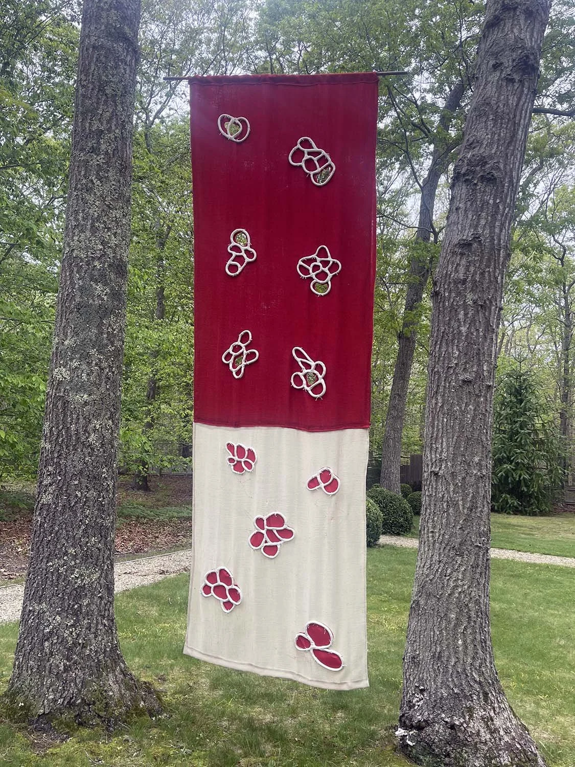 A decorative outdoor textile hanging between two trees in a park or garden. The textile has a color-block design with the top in red and the bottom in off-white. It is decorated with abstract shapes made of white wire or rope, some filled with red, s