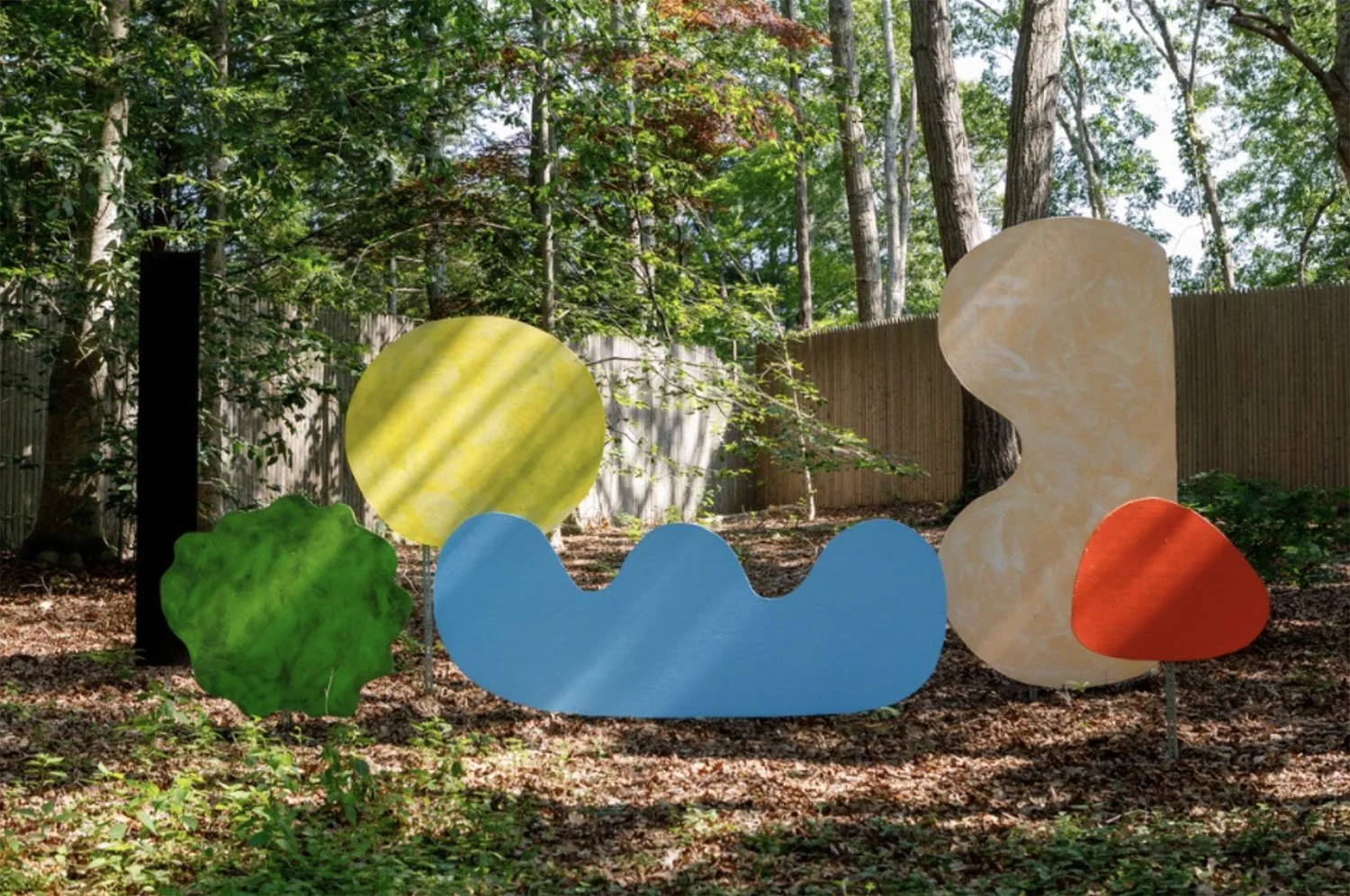 Colorful abstract outdoor art installation in a wooded area, featuring various shapes and colors including a yellow sphere, a green blob, a blue wavy shape, a beige irregular shape, and an orange rounded shape, all mounted on metal stakes.