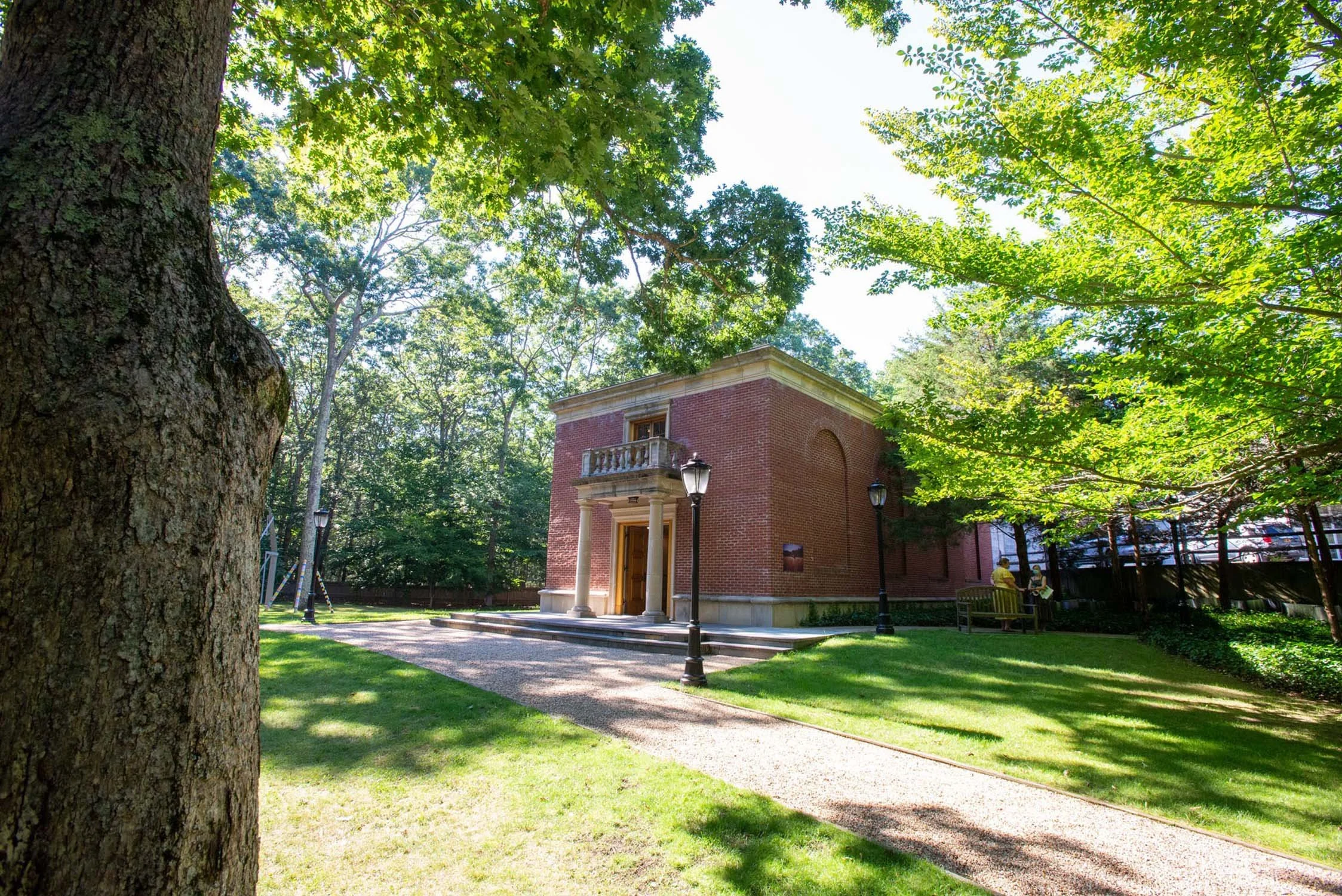 A brick building with a balcony and columns, surrounded by trees and grass, with benches and street lamps nearby.