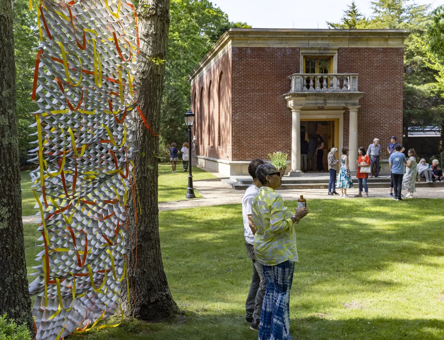 People gathered outdoors near a brick building with columns, trees, and festive decorations, including a hanging paper sculpture with yellow and orange ribbons.