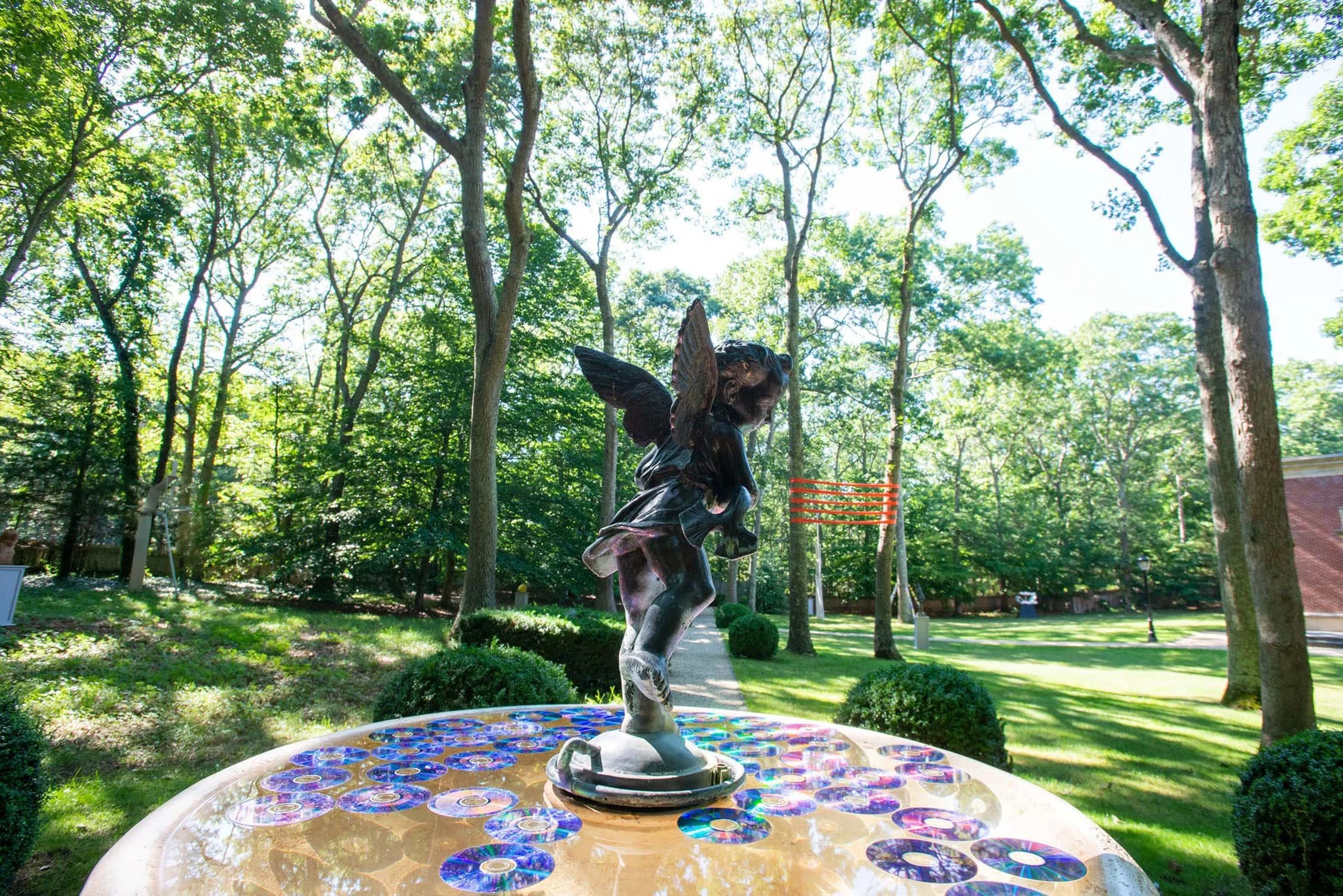 A statue of a fairy on a fountain surrounded by illuminating CD discs, in a green park with trees and grass.