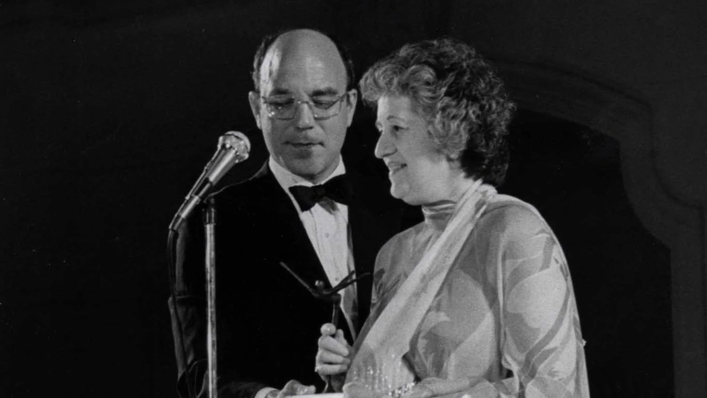 A black and white photograph of Gerson Leiber in a tuxedo with glasses and Judith Leiber in a dress holding glasses, both standing in front of a microphone.