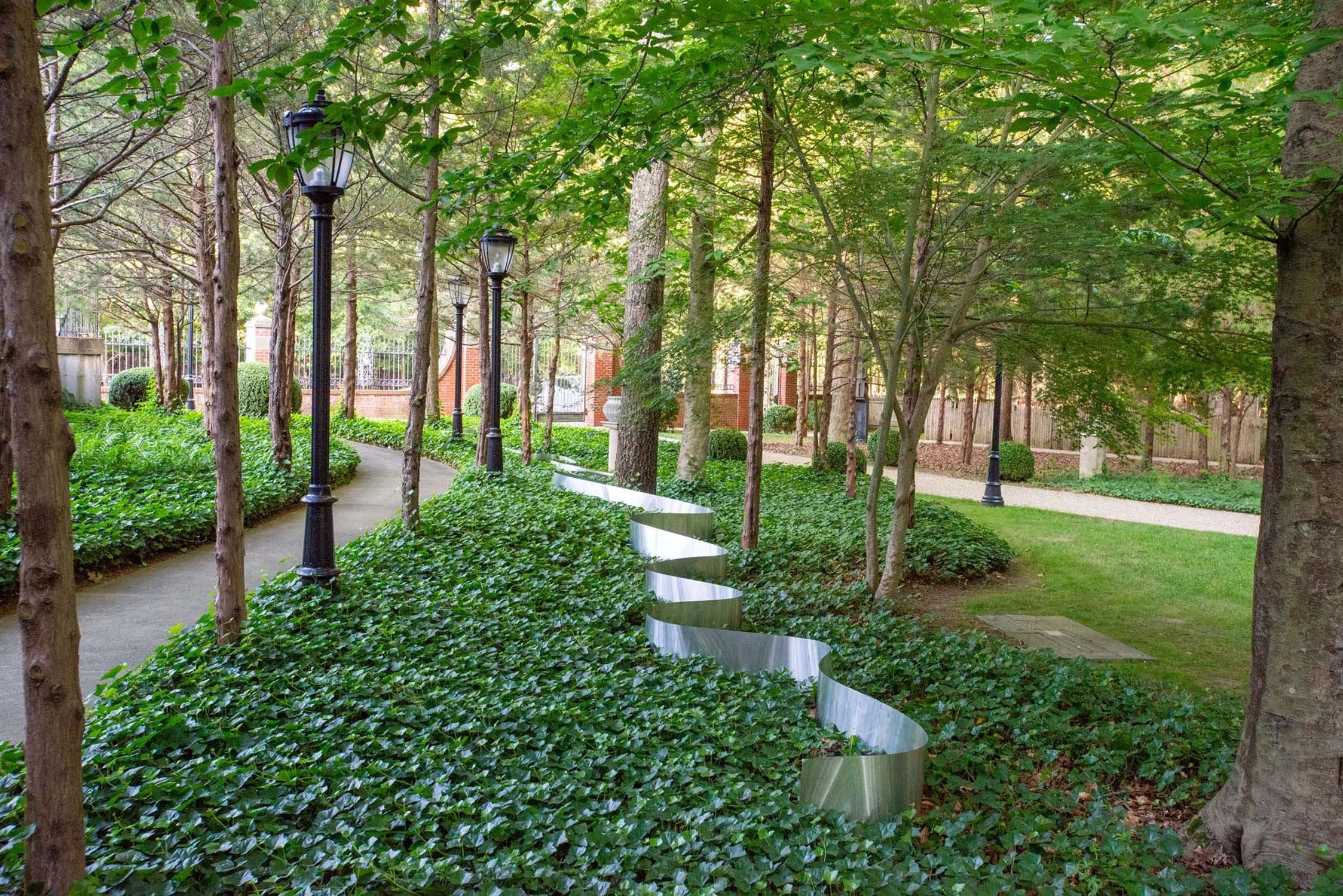A peaceful view of The Leiber Collection garden with a winding pathway lined with black lamp posts, tall trees with green leaves, and lush greenery, including hedges and ground cover, with a modern metallic sculpture running along the ground.